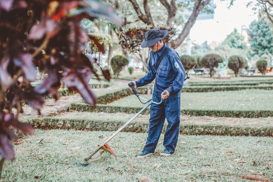 A person dressed in a black t-shirt, black and white striped athletic pants, and bright green waterproof boots is crouched down on a garden pathway, carefully removing a weed from the soil with a smal